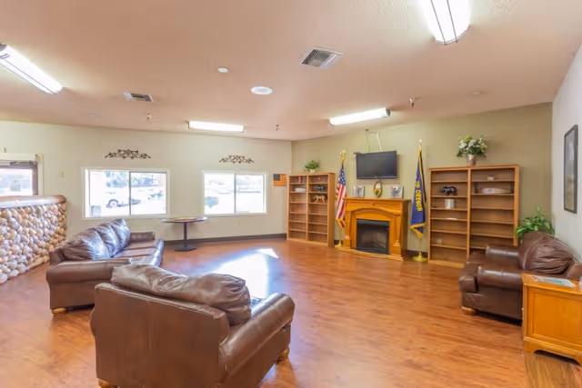 A spacious common area with wooden flooring, three brown leather sofas arranged around the room, a wooden fireplace with a TV mounted above it, two wooden bookshelves on either side of the fireplace, and two flags placed next to the fireplace. The room has two windows letting in natural light and a stone half-wall on the left side.