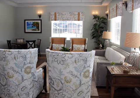 A cozy living room with floral patterned armchairs in the foreground, two wooden chairs with cushions near a window in the background, a gray sofa on the right side, a wooden side table with a lamp and decorative items, a tall green plant, and framed artwork on the wall.