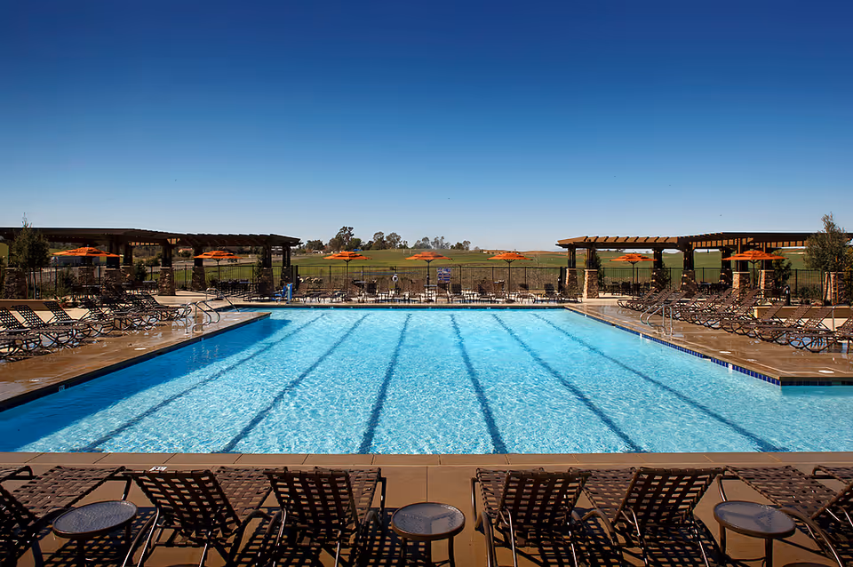 Outdoor swimming pool with clear blue water surrounded by lounge chairs and tables. There are orange umbrellas and pergolas on the far side of the pool, with a green field and trees in the background under a clear blue sky.