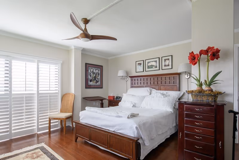 Sunlit bedroom with a wooden bed dressed in white linens, matching nightstands and dresser, plantation shutters, and a ceiling fan.