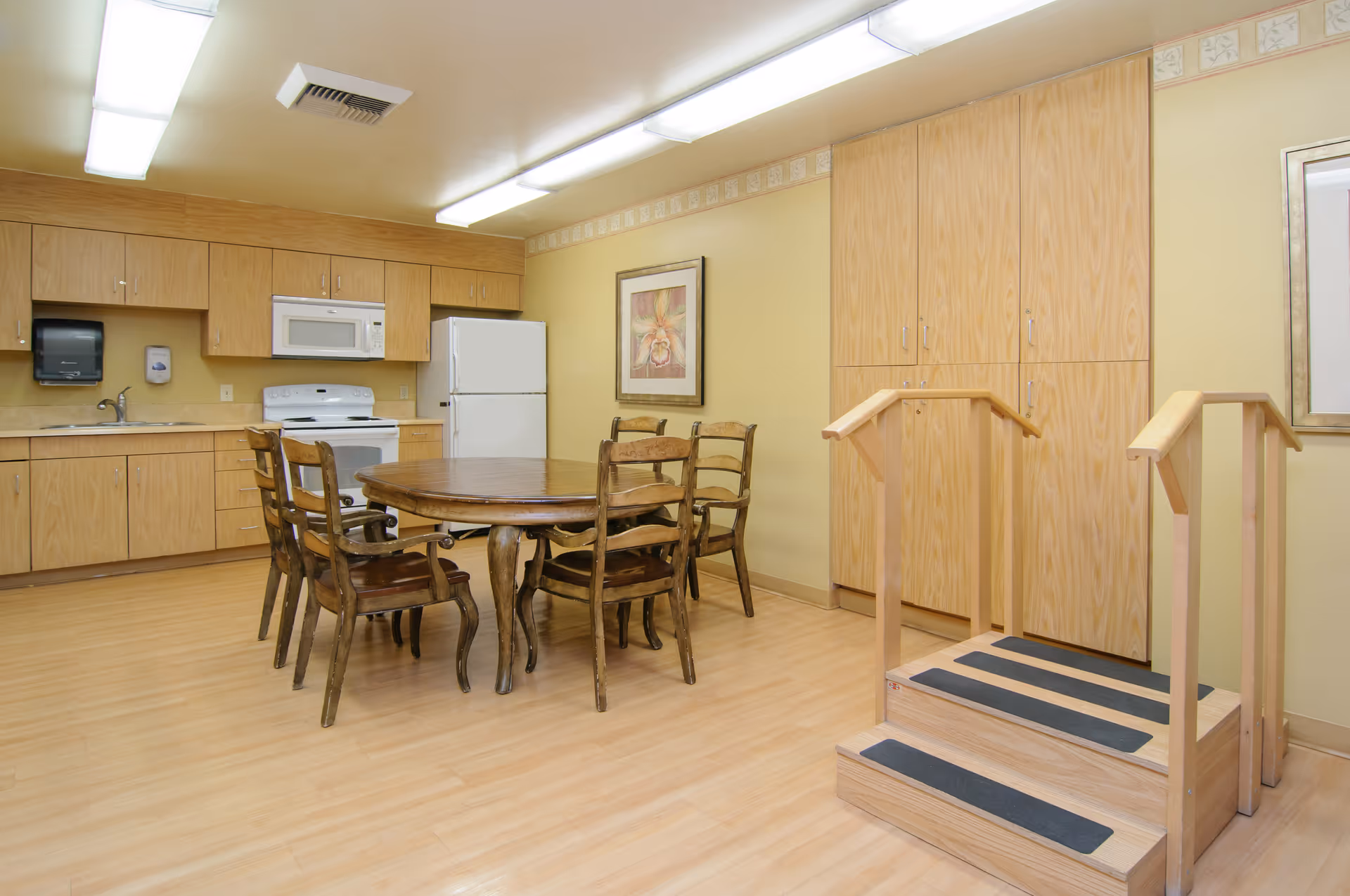 Communal kitchenette and dining area with a round wooden table and chairs, appliances and cabinets, and a small wooden practice staircase.