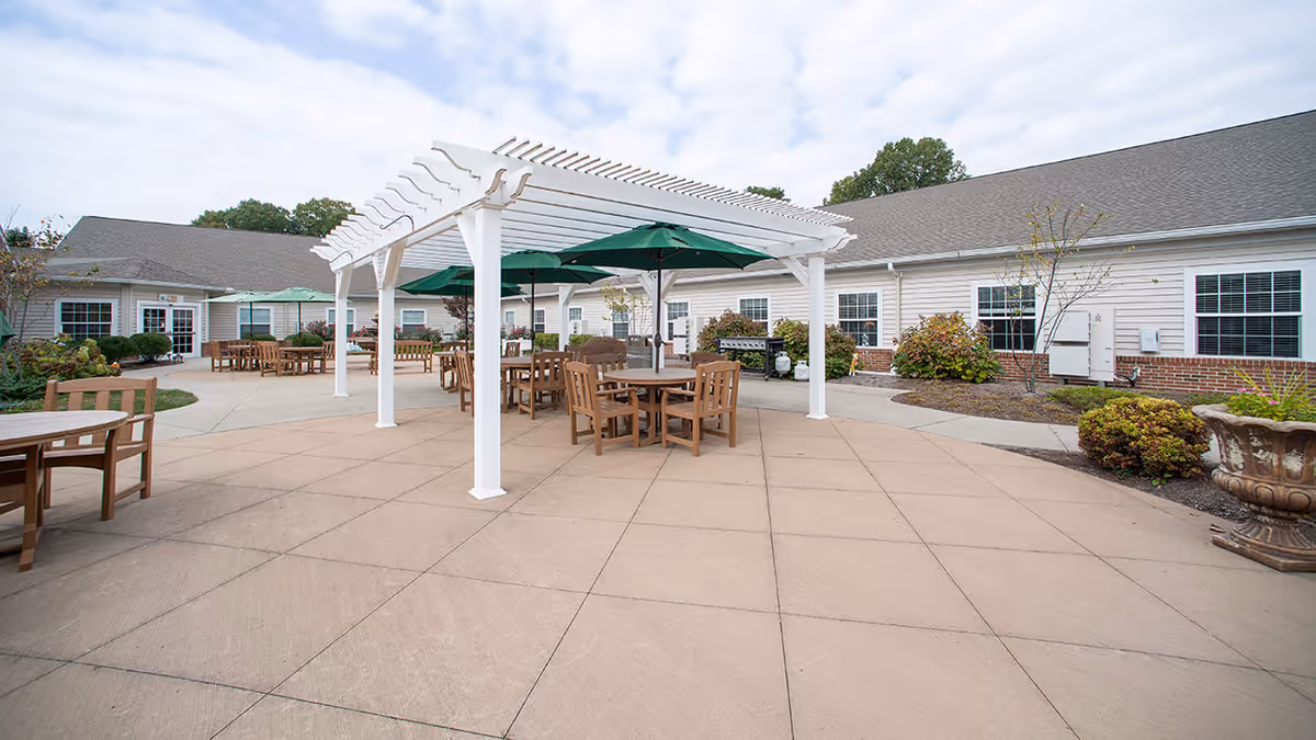 Outdoor patio area at Danbury Massillon with wooden tables and chairs under green umbrellas and a white pergola. The patio is surrounded by a single-story building with white siding and brick accents, and there are some small bushes and plants along the building.
