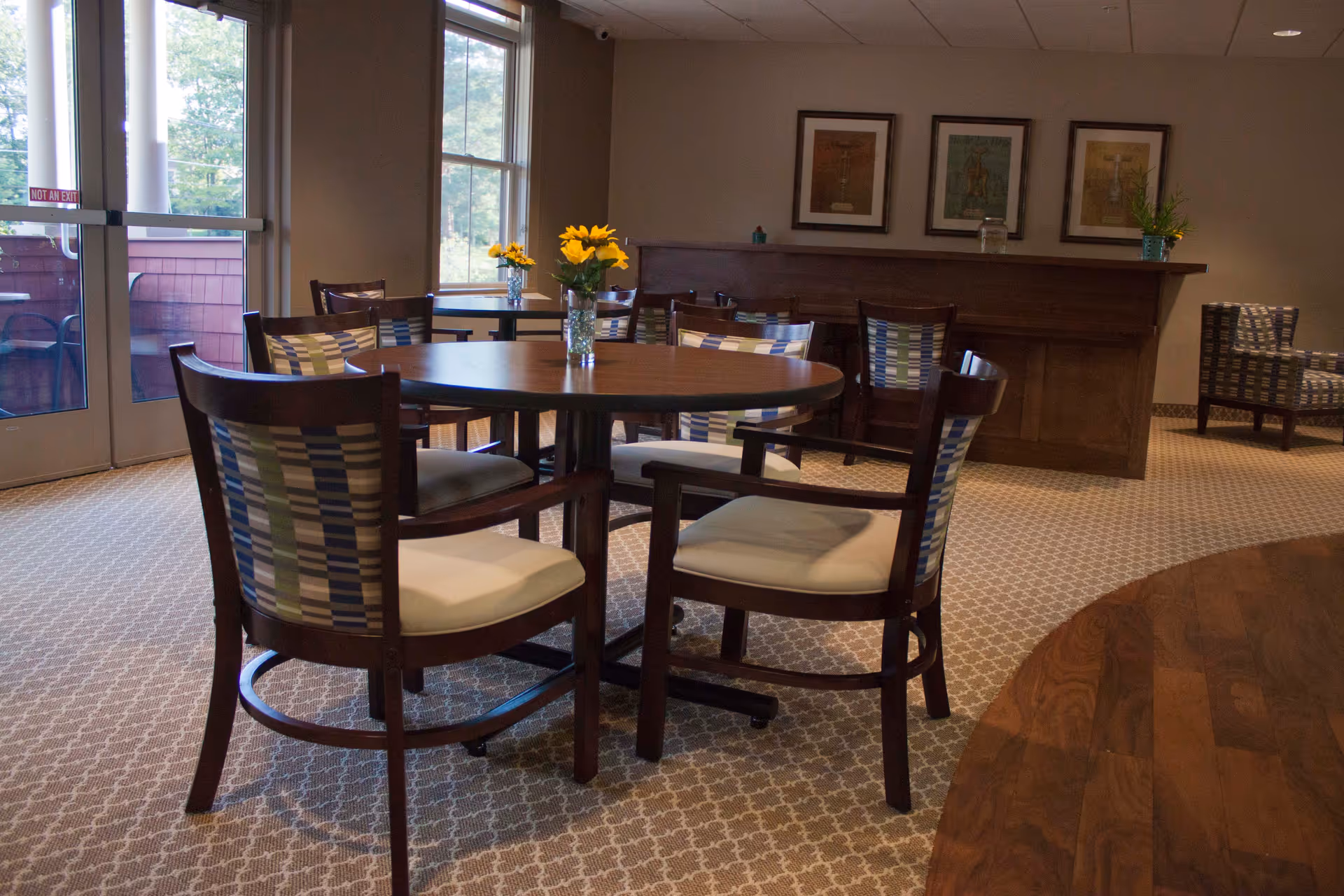 A cozy senior living common area with round wooden tables and cushioned chairs featuring patterned backs. Sunflowers in glass vases decorate the tables. Large windows and glass doors let in natural light, and framed artwork hangs on the beige walls. A wooden counter and additional seating are visible in the background.