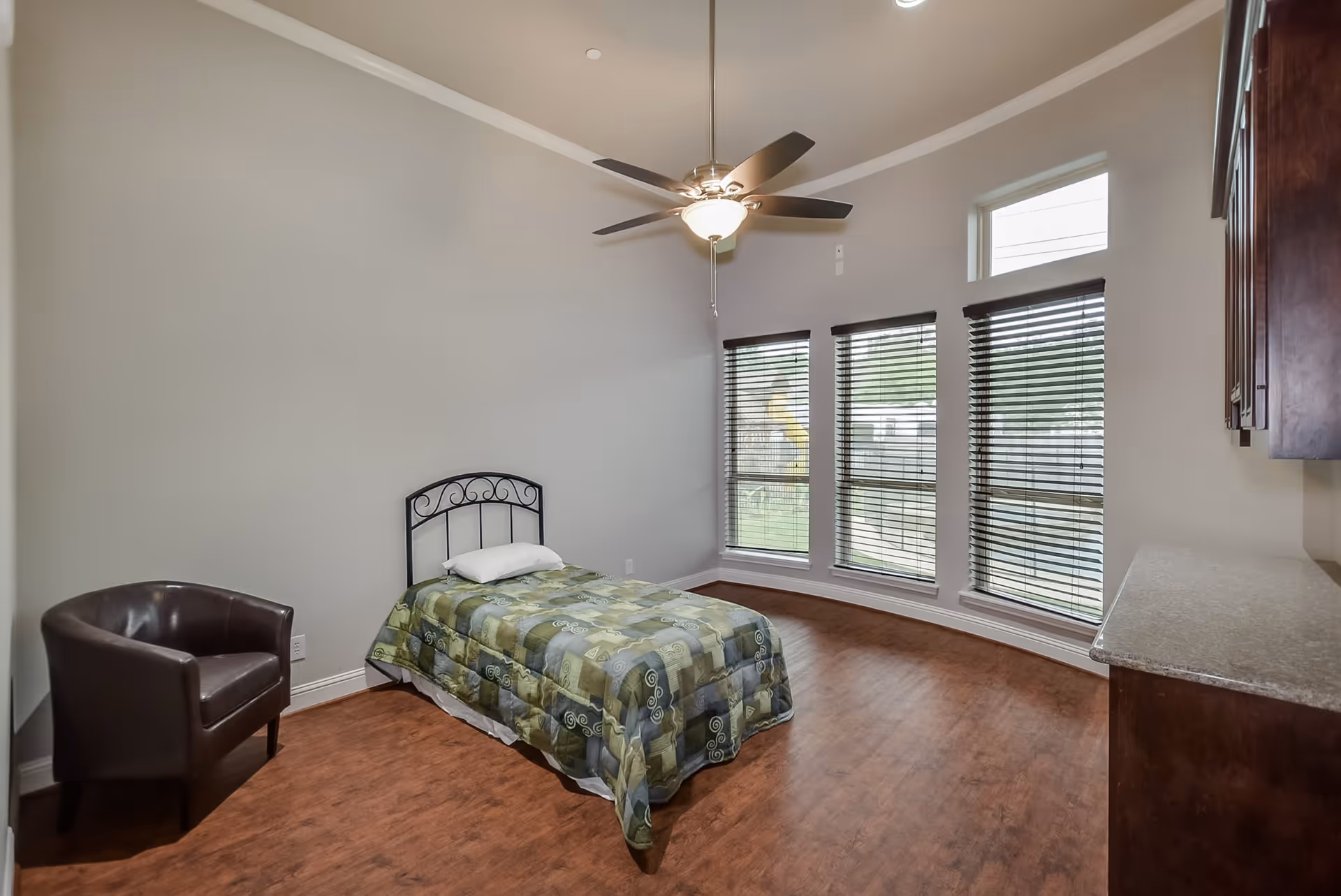 A simple bedroom with a single bed covered with a green patterned comforter and a white pillow. There is a dark brown armchair to the left of the bed. The room has wooden flooring, three large windows with blinds, and a ceiling fan with a light fixture. On the right side, there is a countertop and dark wooden cabinets mounted on the wall.