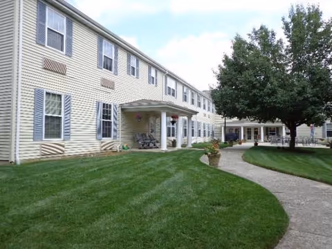 Two-story beige building with blue shutters, curved walkway, porch seating, and a manicured lawn.