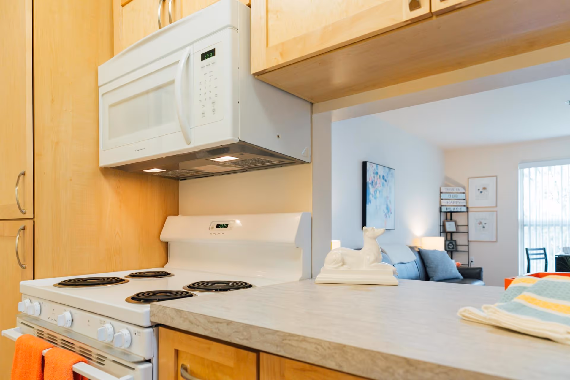 A kitchen area featuring a white electric stove with four coil burners and an orange towel hanging on the oven handle. Above the stove is a white microwave mounted under wooden cabinets. A countertop extends from the stove area, with a white decorative dog figurine and a folded striped towel on it. In the background, a living room area is visible with a couch, wall art, a lamp, and a window with vertical blinds.