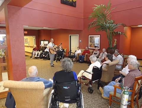 A group of elderly residents seated in a common area of an assisted living facility, some in wheelchairs and others in chairs, attentively watching a man playing an acoustic guitar. The room has red walls, a large potted plant, and framed artwork on the walls.