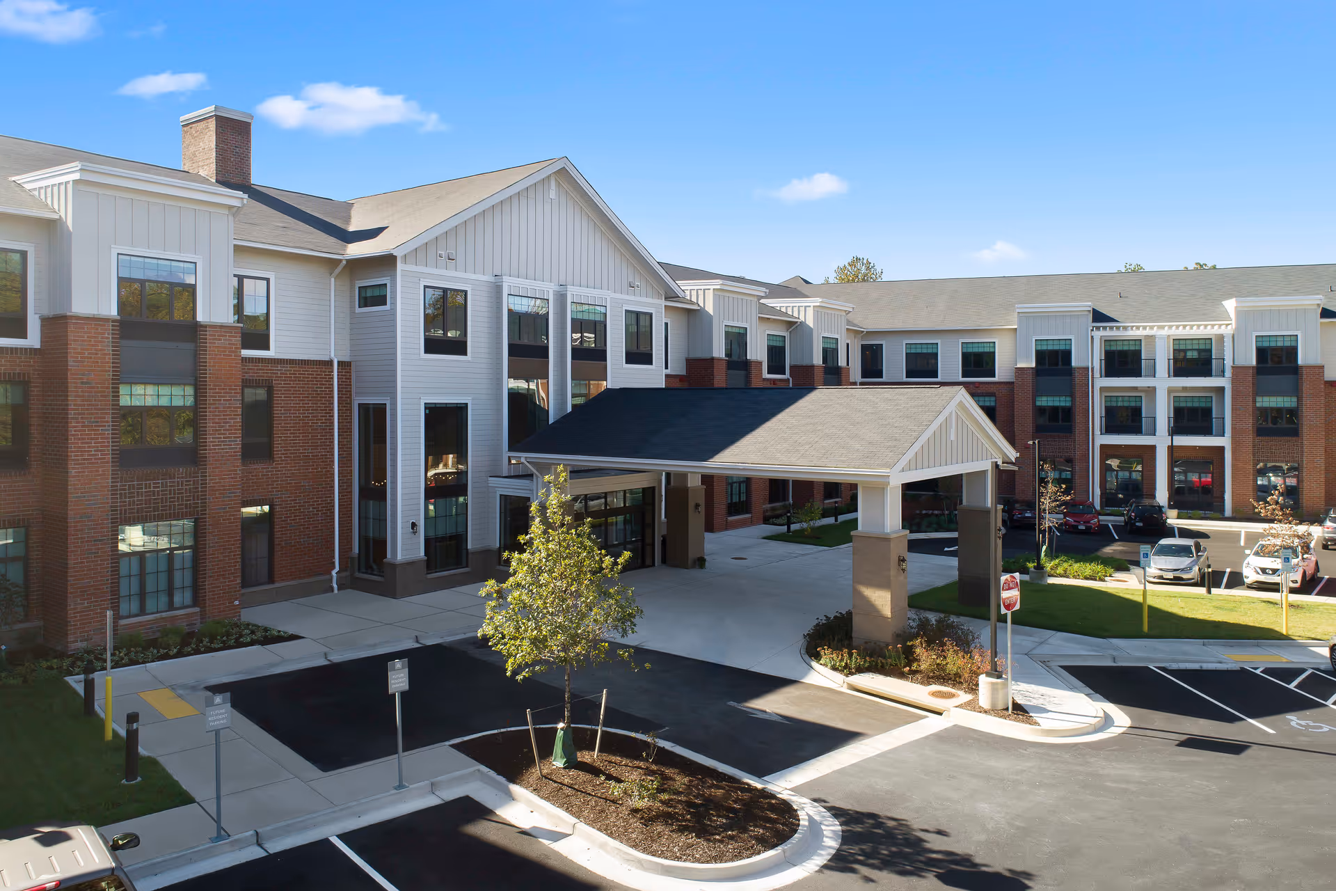Exterior view of a senior living facility named Elancé at Tuckahoe, featuring a large three-story building with a covered entrance driveway, brick and white siding facade, multiple windows, a small landscaped area with a tree, and a parking lot with several cars.