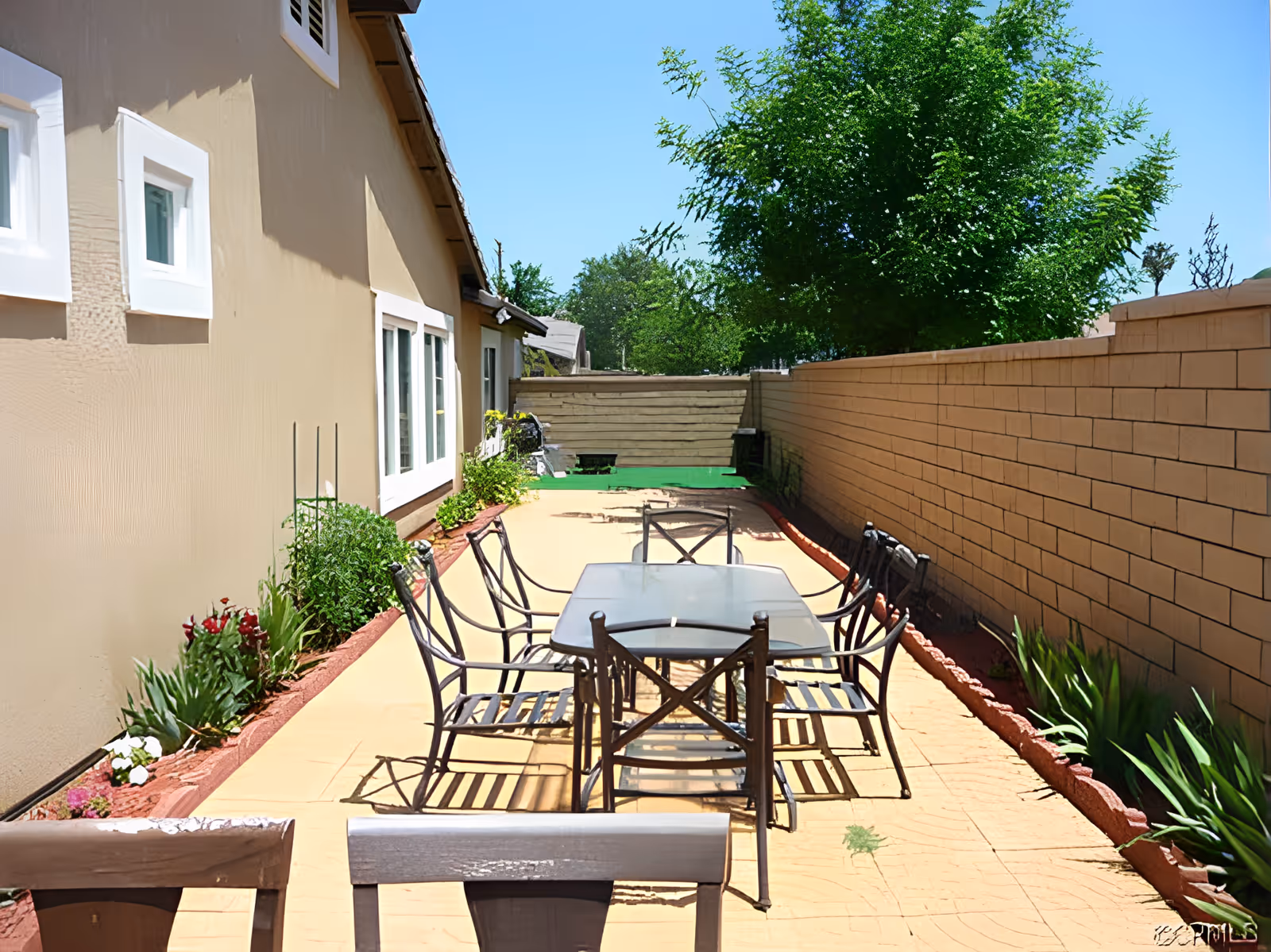 Outdoor patio area with a rectangular glass table and six metal chairs arranged around it. The patio is bordered by a beige building wall on the left and a brick wall on the right, with flower beds containing green plants and flowers along both sides. Trees and a clear blue sky are visible in the background.