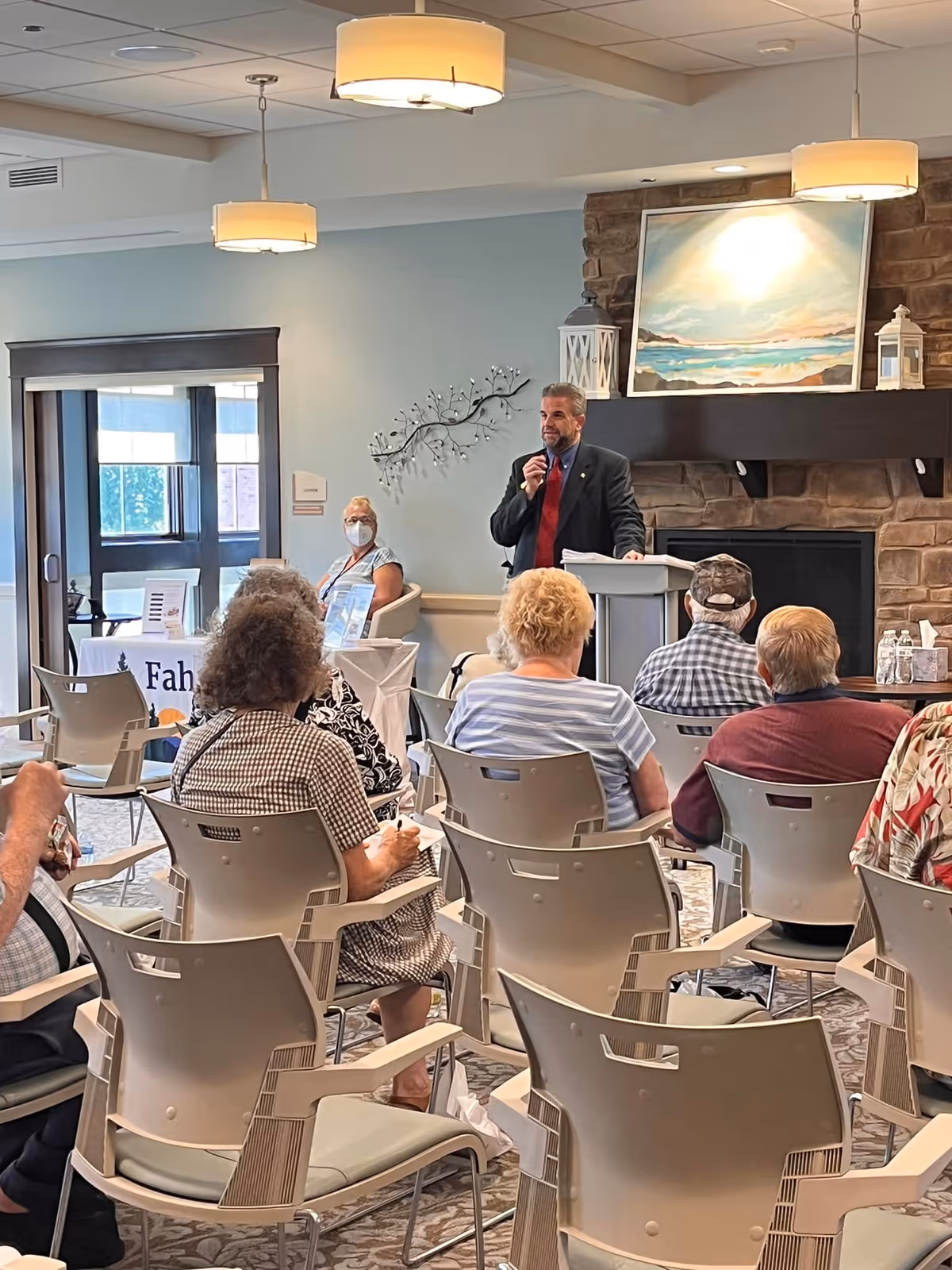 A man in a suit and red tie is speaking at a podium in a room with a stone fireplace and a large painting above it. Several elderly people are seated in chairs facing him, listening attentively. One woman in the background is wearing a face mask. The room has soft lighting with hanging lamps and a calm, welcoming atmosphere.