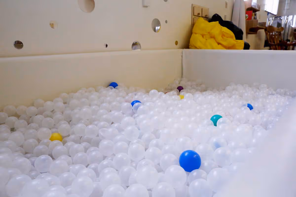 A large indoor ball pit filled mostly with white plastic balls and a few colored balls including blue, yellow, purple, and teal. The ball pit has white padded walls with circular cutouts. In the background, there is a yellow bean bag chair and some wooden furniture near windows letting in natural light.