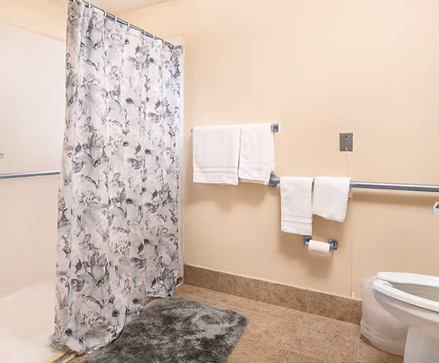 Bathroom with a floral shower curtain, beige tiled floor, two white towels hanging on a towel rack, a toilet with a plastic cover, and a gray bath mat on the floor.
