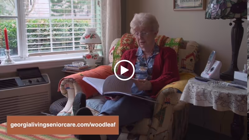 An elderly woman with glasses and a red cardigan is sitting in a cozy armchair with a colorful quilt, reading a book. The room has a window with blinds, a small table with a lace tablecloth, a cordless phone, a lamp with a stained glass shade, and an air conditioning unit below the window.