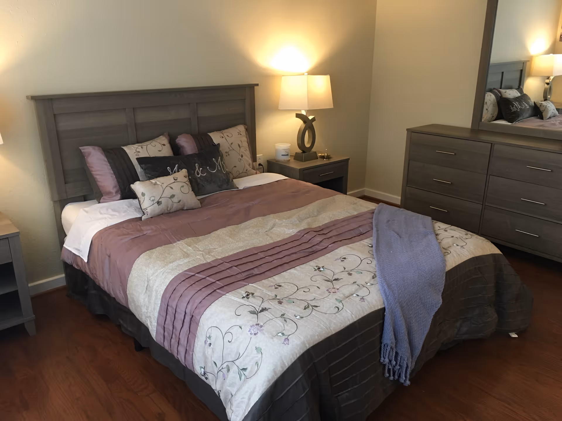 A neatly made bed with a wooden headboard in a bedroom. The bed has a floral and striped comforter in shades of purple, beige, and gray, with multiple decorative pillows including one with the text 'Mr. & Mrs.'. There is a wooden nightstand with a modern lamp on the right side of the bed, and a matching wooden dresser with a large mirror on the right wall. The room has wooden flooring and soft lighting.