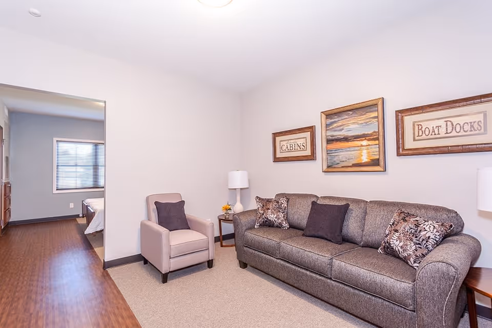 A cozy living room area with a gray upholstered sofa adorned with patterned and solid dark pillows, a beige armchair with a dark pillow, a side table with a white lamp and a small flower arrangement. On the wall above the sofa are three framed pictures, one depicting a sunset over water and two with text reading 'CABINS' and 'BOAT DOCKS'. The room has light-colored walls and carpeted flooring, with a view into an adjacent bedroom featuring a bed and window with blinds.