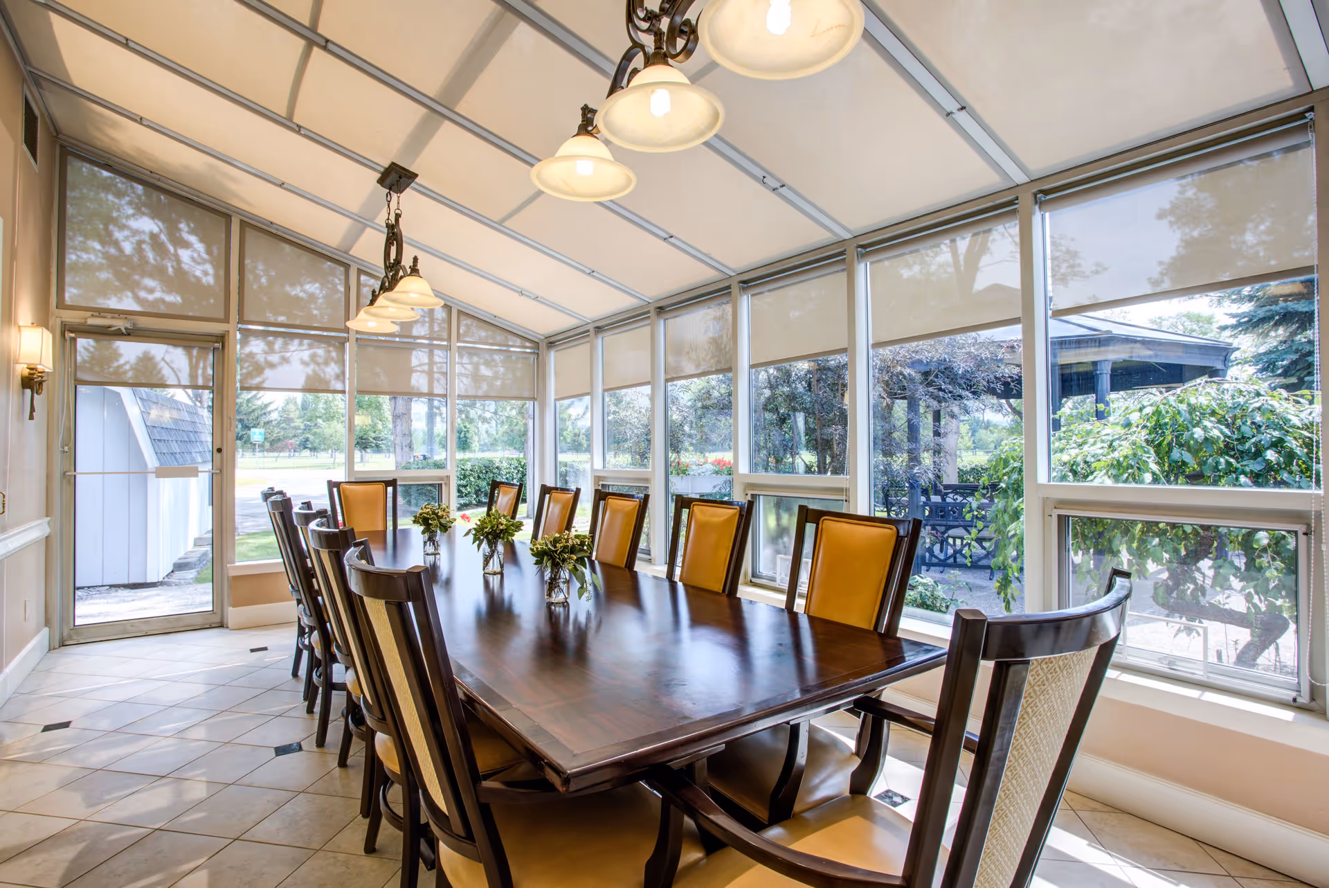 A bright dining room with a long wooden table surrounded by twelve chairs with yellow cushions. The room has large windows on three sides with beige roller shades, allowing natural light to fill the space. There are three small flower arrangements on the table and a chandelier with five lights hanging above it. Outside the windows, greenery and trees are visible.