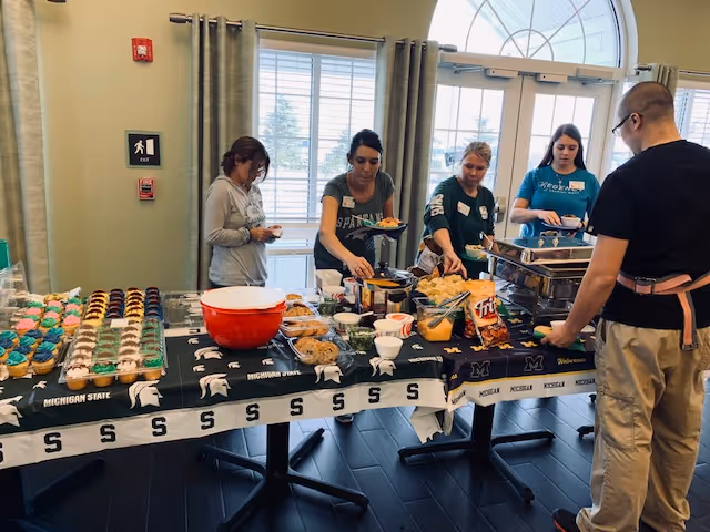 Residents and staff serve themselves from a buffet table with cupcakes, snacks, and hot dishes in a dining area.