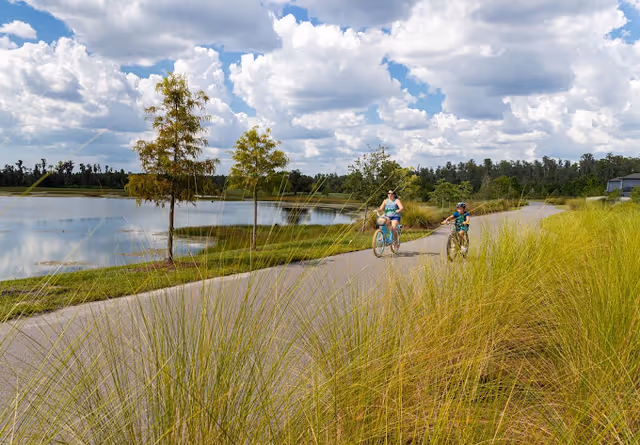Two cyclists ride along a paved path beside a lake with tall grasses, trees, and a partly cloudy sky.