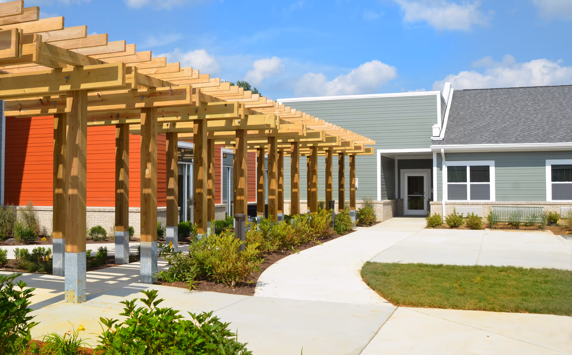 Outdoor view of a senior living facility showing a wooden pergola structure casting shadows on a concrete walkway, with green shrubs and grass surrounding the path. The building has a combination of red and gray siding with white trim and multiple windows under a partly cloudy blue sky.