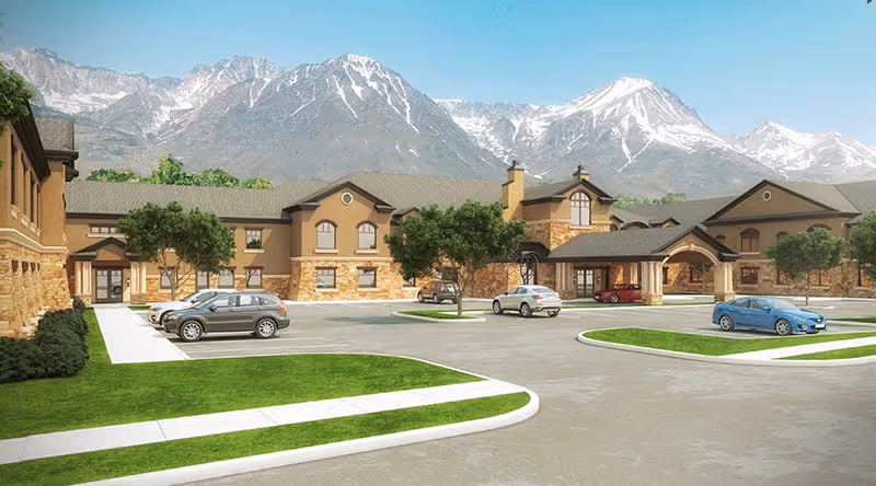 Exterior view of The Ridge Cottonwood senior living facility with a parking lot in front, several cars parked, green lawns, and large mountains in the background under a clear blue sky.