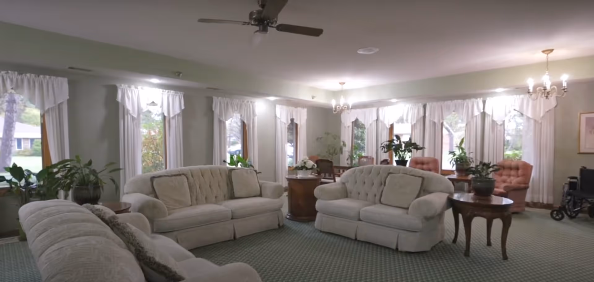 A spacious living room with multiple beige upholstered sofas and armchairs arranged around wooden side tables with potted plants. The room has large windows with white curtains, green carpet flooring, and ceiling lights including chandeliers and a ceiling fan. There is a wheelchair visible in the corner near the windows.