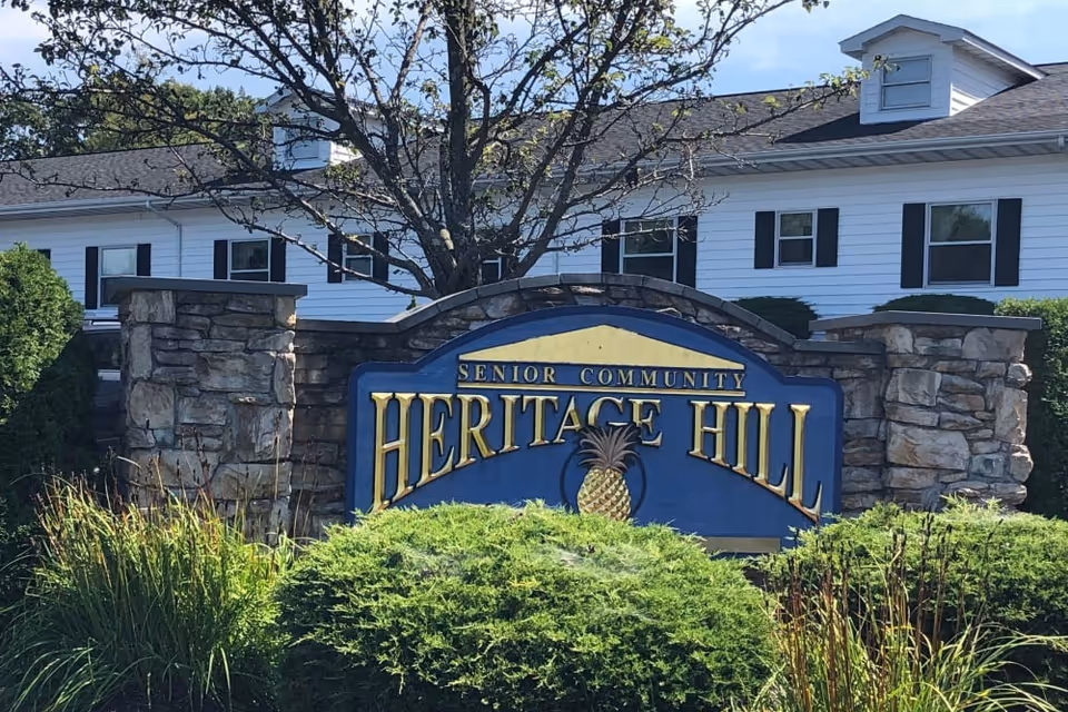 Stone sign with the text 'Senior Community Heritage Hill' in front of a white building with black shutters and a tree behind the sign, surrounded by green bushes and grass.