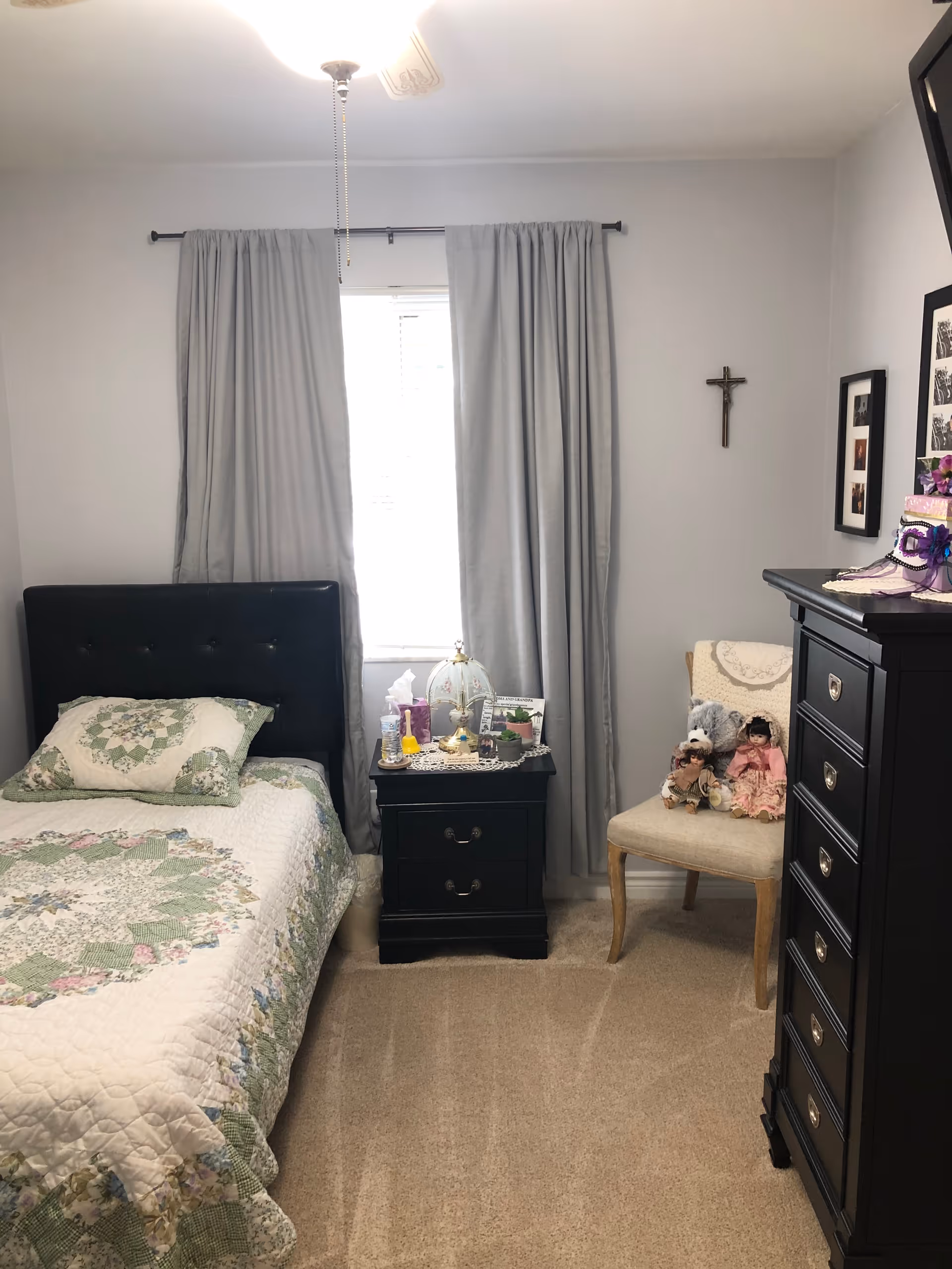 A cozy bedroom with a single bed covered in a quilted bedspread with green and pink patterns. Next to the bed is a black nightstand with various items including a lamp, tissues, and personal care products. A window with light gray curtains is behind the nightstand. To the right, there is a beige chair with dolls and a teddy bear on it, and a black dresser with drawers. A crucifix hangs on the wall above the chair, and framed pictures are partially visible on the wall.