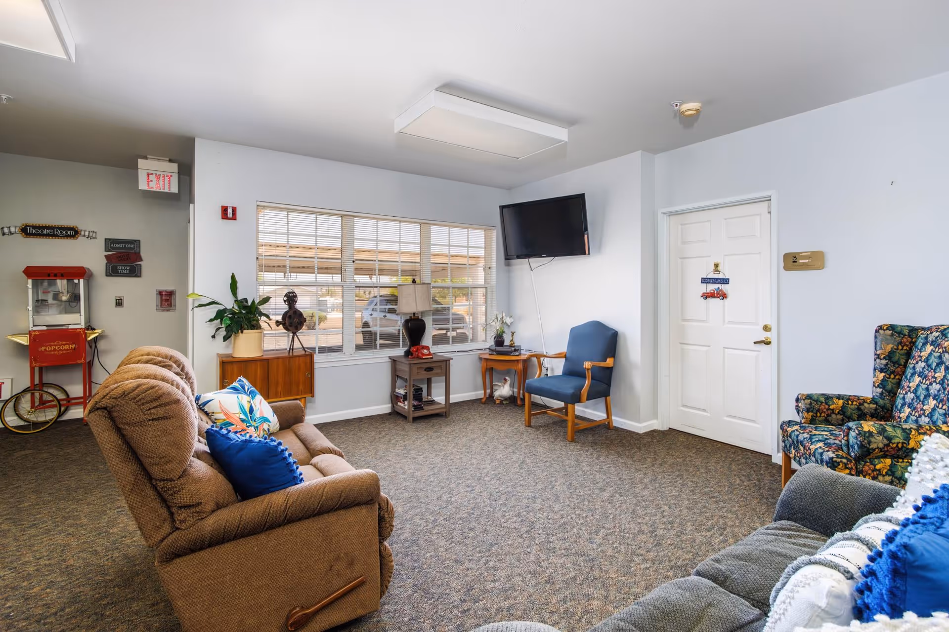 A cozy living room area in an assisted living facility with several upholstered chairs and sofas arranged around the room. A flat-screen TV is mounted on the wall, and there are decorative items including a lamp, potted plant, and a vintage popcorn machine near the exit door. Large windows allow natural light to fill the space.