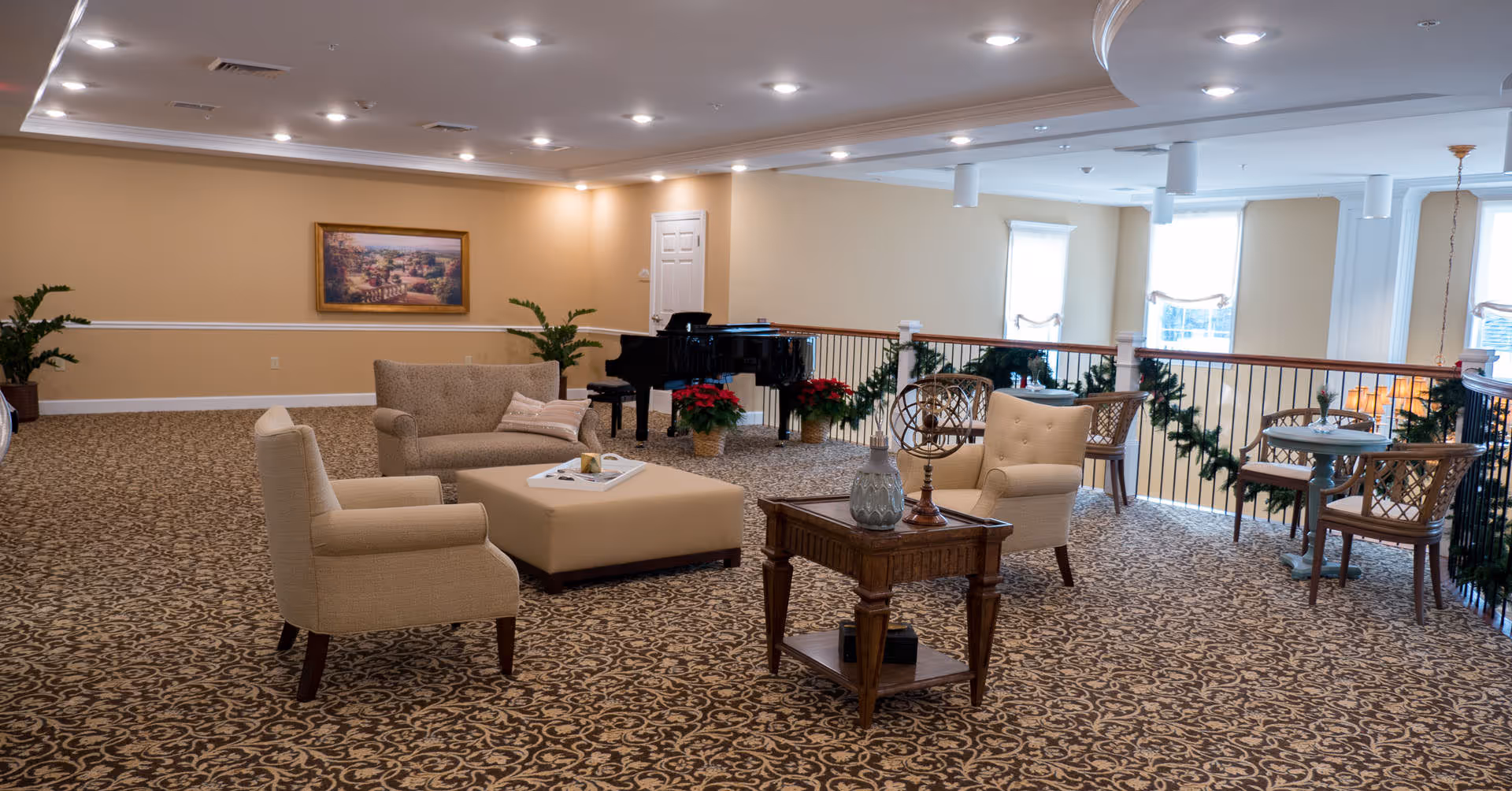 Bright, carpeted communal lounge with upholstered chairs and tables, a grand piano, and garland-decorated railing overlooking windows.