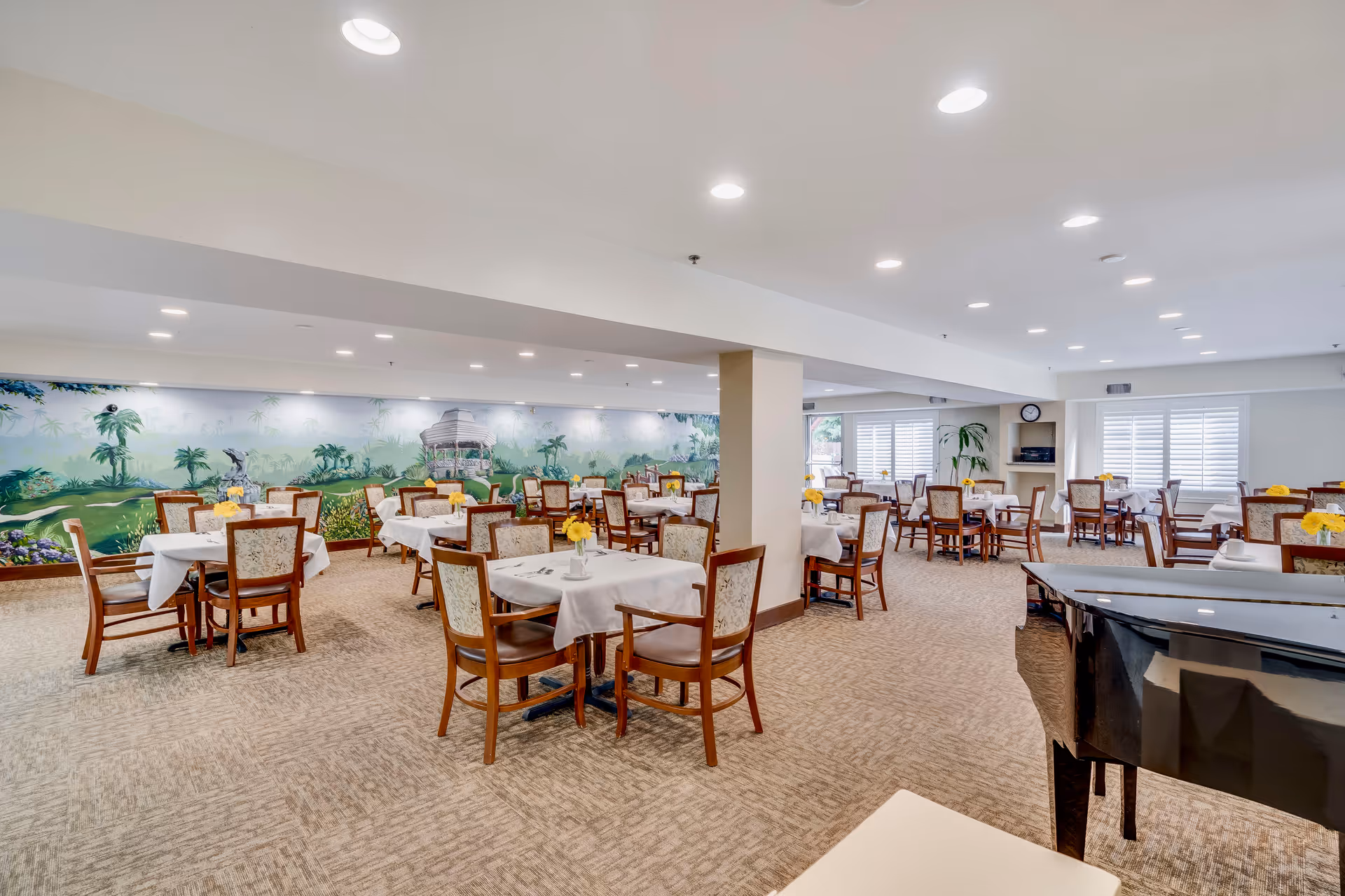 A spacious dining room with multiple tables covered in white tablecloths, each set with chairs and small yellow flower centerpieces. The room has beige carpeting, bright overhead lighting, and a large mural depicting a tropical scene on one wall. A black grand piano is visible in the foreground on the right side.