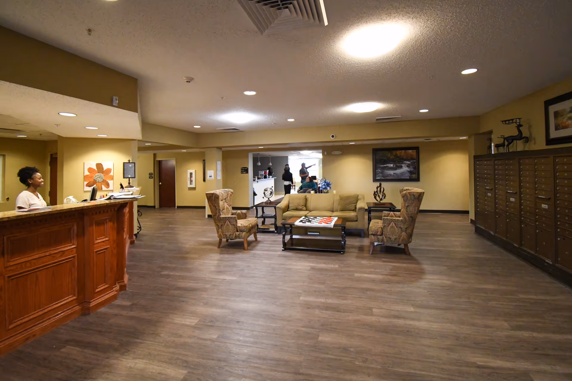 Interior view of a senior living facility lobby with a wooden reception desk on the left where a woman is seated. In the center, there is a seating area with a beige sofa, two patterned armchairs, and a coffee table with a checkerboard game. On the right side, there are multiple mailboxes along the wall. The background shows a hallway with three people standing and talking. The walls are painted beige and the floor has wood-like flooring.