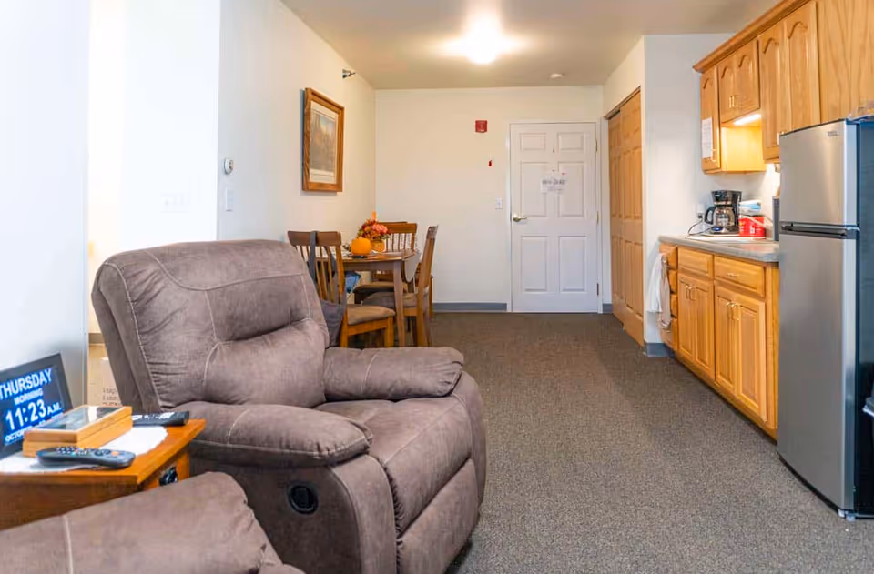 Interior view of a senior living facility room showing a brown recliner chair in the foreground, a small wooden side table with a digital clock and remote controls, a dining table with four chairs and a floral centerpiece, and a kitchenette area with wooden cabinets, a coffee maker, and a stainless steel refrigerator. The room has carpeted flooring and white walls with a framed picture hanging.
