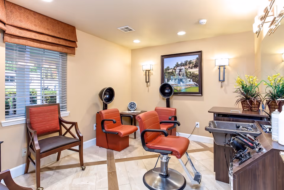 Interior of a hair salon area in a senior living facility with two red salon chairs, two black hair dryers, a wooden chair with red cushion, a small table with a clock, a large mirror, and decorative plants. The walls are painted beige and there is a window with blinds and a brown valance.