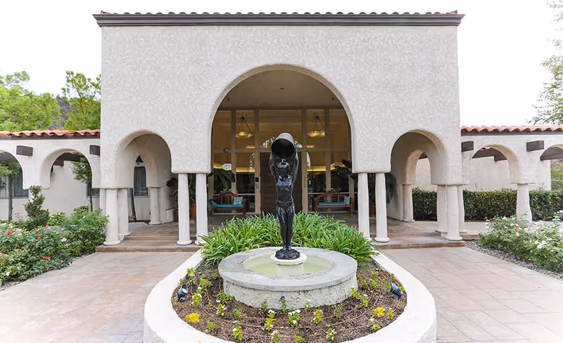 Exterior view of Silverado Calabasas Memory Care Community featuring a central fountain with a statue of a child holding a large bowl above their head, surrounded by a circular flower bed. The building has a stucco finish with arched walkways and red tile roofing. Green shrubs and trees are visible around the entrance area.