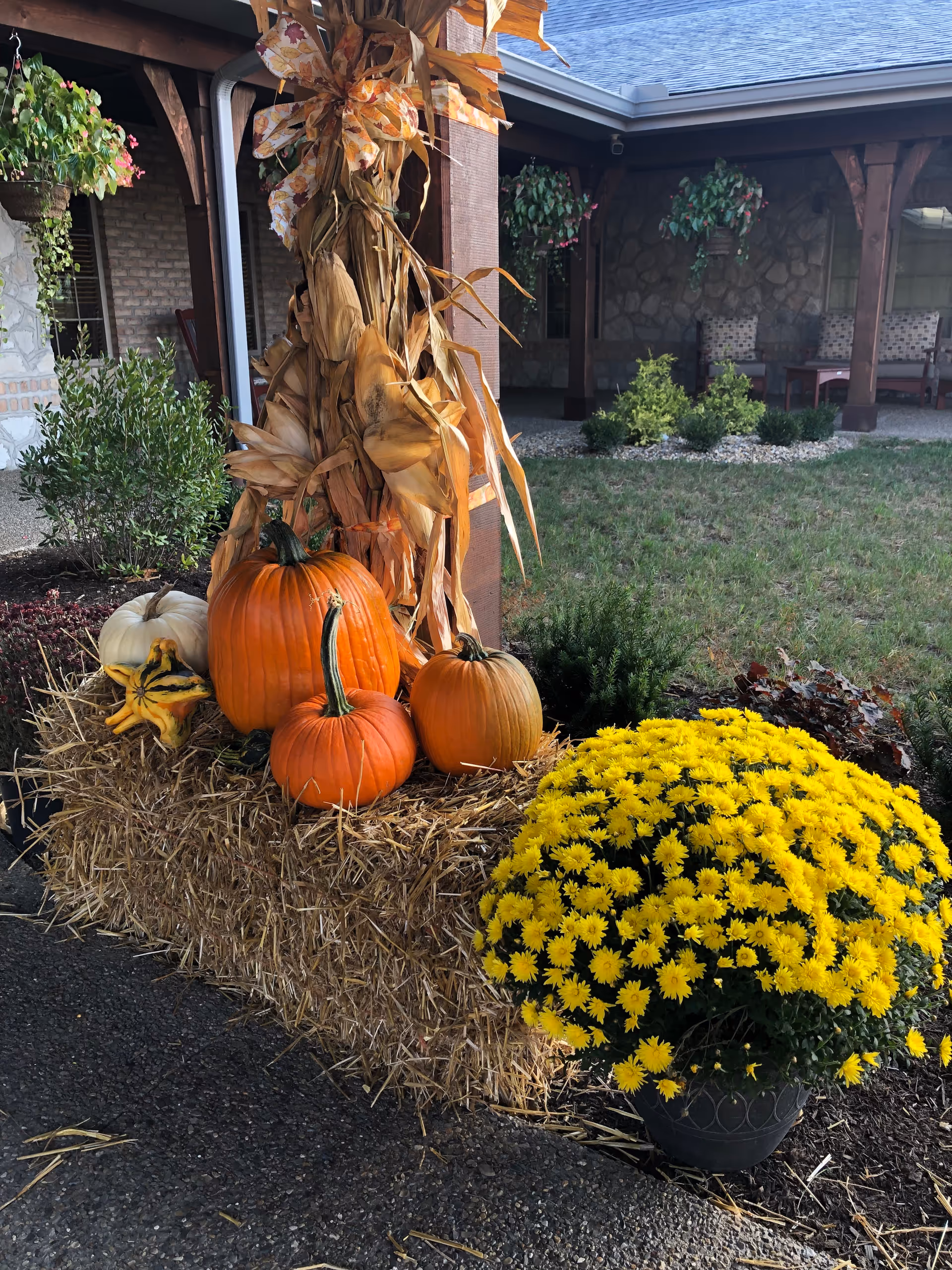 A fall-themed outdoor decoration featuring several pumpkins of different sizes and colors arranged on a bale of hay, with dried corn stalks tied to a wooden post behind them. To the right, there is a large pot of bright yellow chrysanthemums. In the background, there is a covered patio area with chairs and hanging plants.