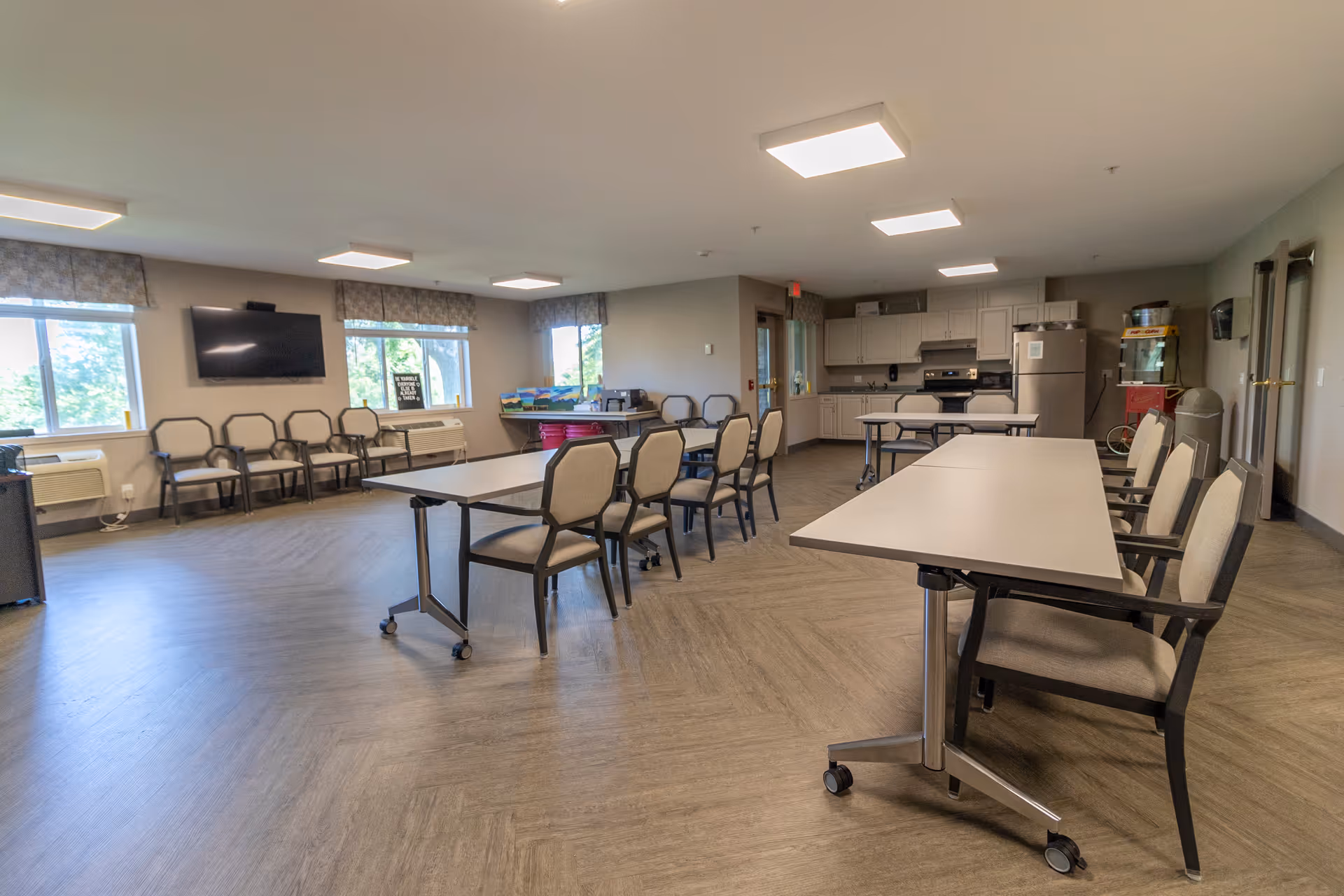 Spacious community dining/activity room with folding tables and chairs, a kitchenette in the back, and a wall-mounted TV.