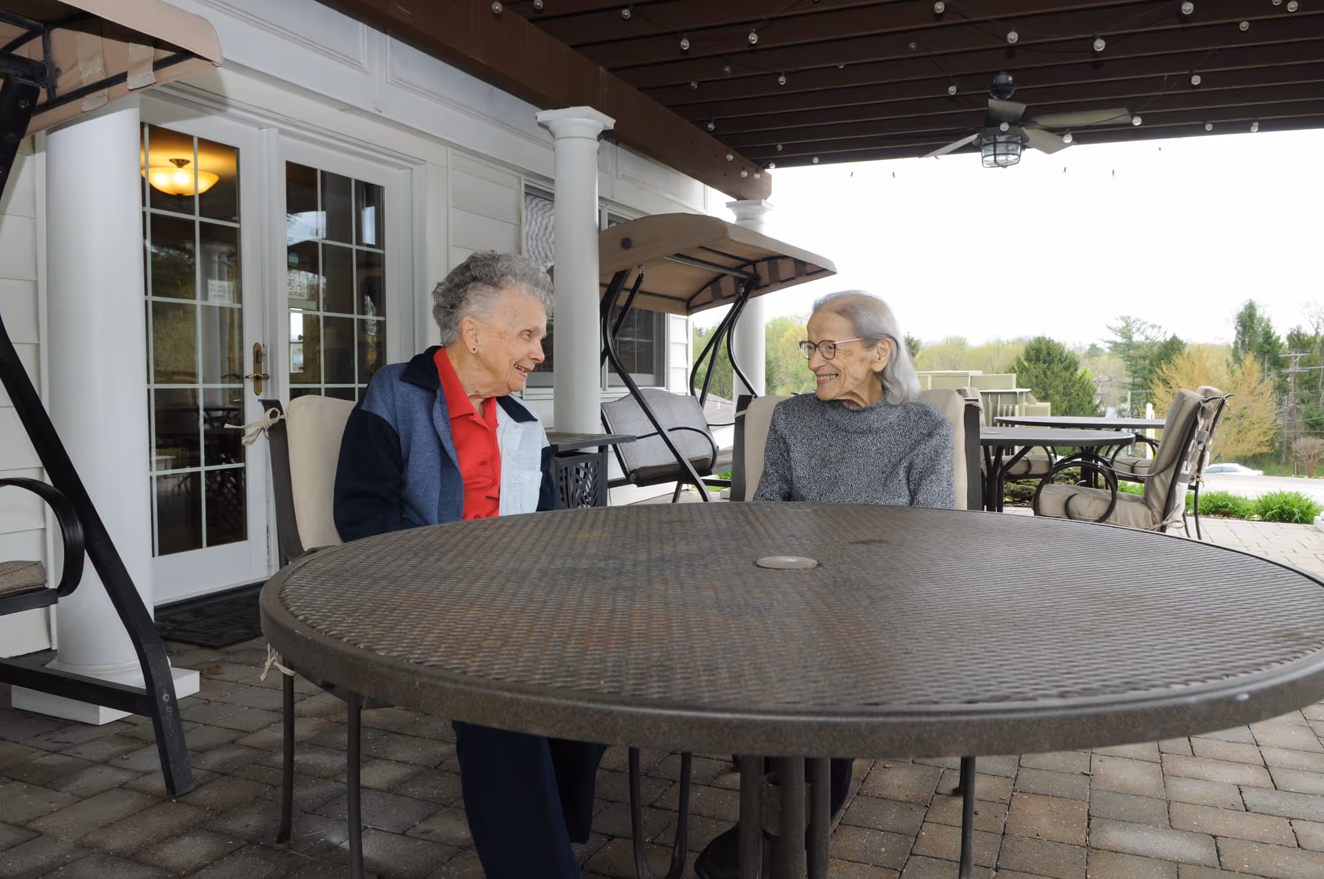 Two elderly women sitting and smiling at each other on a covered outdoor patio with a round metal table in front of them. The patio has cushioned chairs, a swing, and a view of trees and greenery in the background.