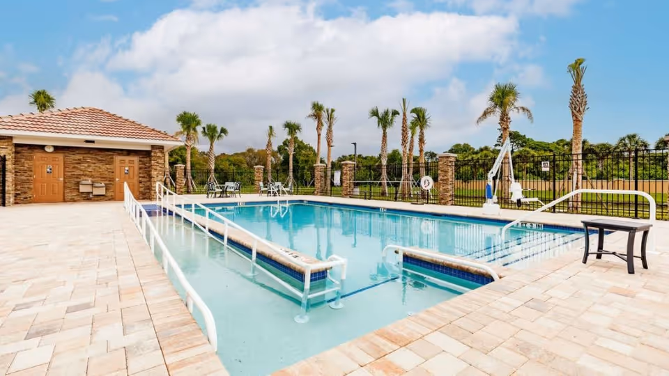 Outdoor swimming pool with handrails, seating, palm trees, and a small pool house under a partly cloudy sky.