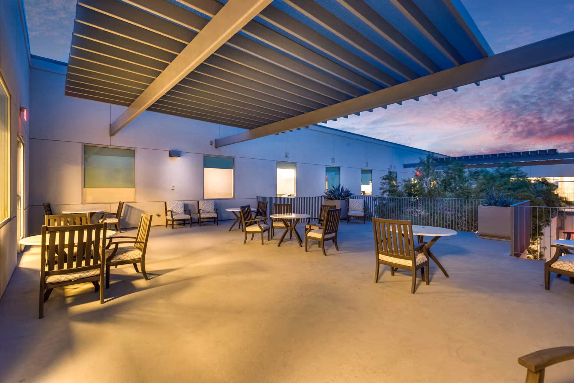 Outdoor patio area at dusk with several round tables and cushioned wooden chairs arranged under a large pergola attached to a building. The sky is colorful with a mix of blue and pink hues, and there are plants and trees visible in the background.