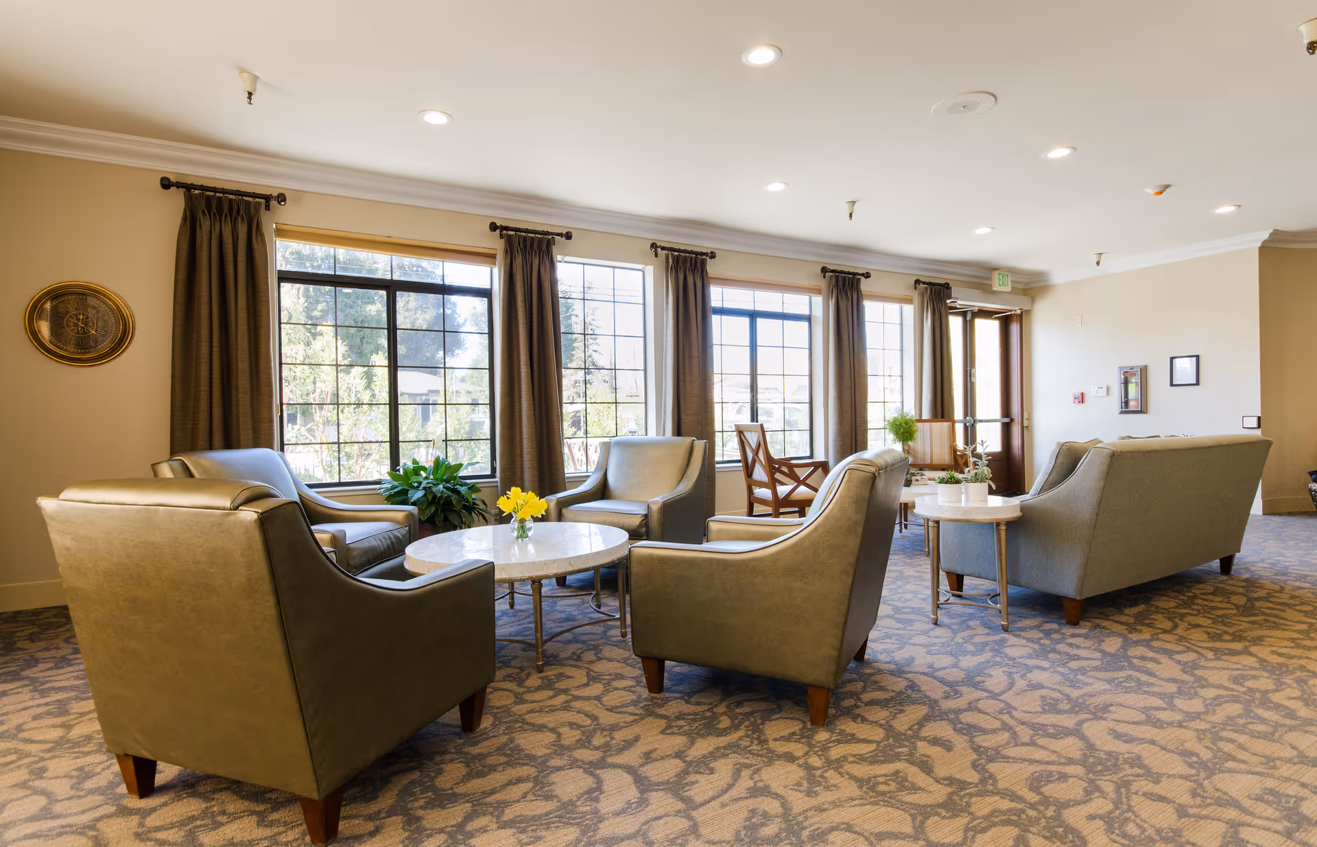 Sunlit assisted living lounge with upholstered armchairs and sofas grouped around round coffee tables by large windows.