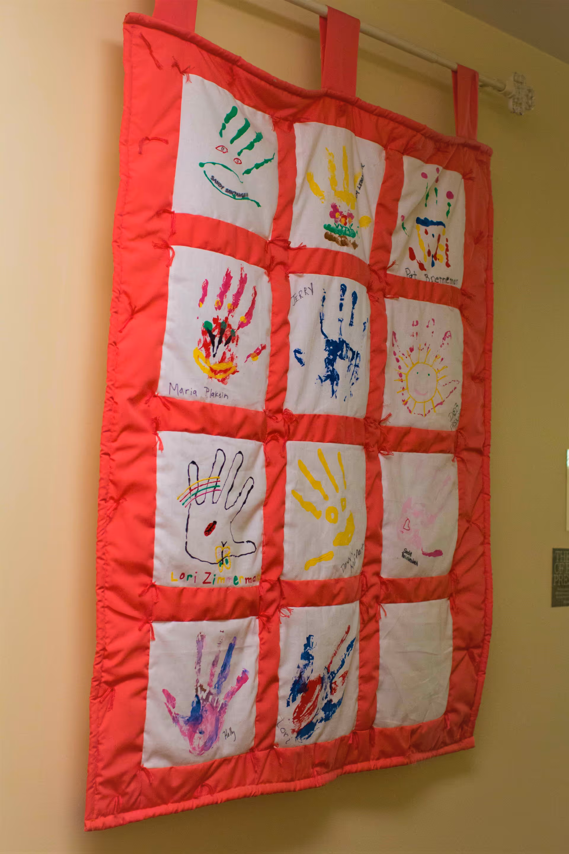 Colorful quilted wall hanging with painted handprint squares displayed on a rod against a beige wall.