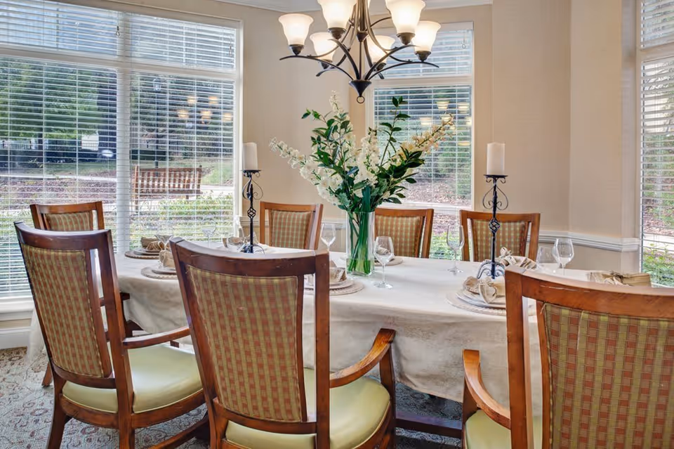 A dining room with a rectangular table covered with a white tablecloth, set with plates, napkins, wine glasses, and two candle holders. There is a vase with white flowers in the center of the table. The room has large windows with white blinds, allowing natural light to brighten the space. Wooden chairs with patterned upholstery surround the table.