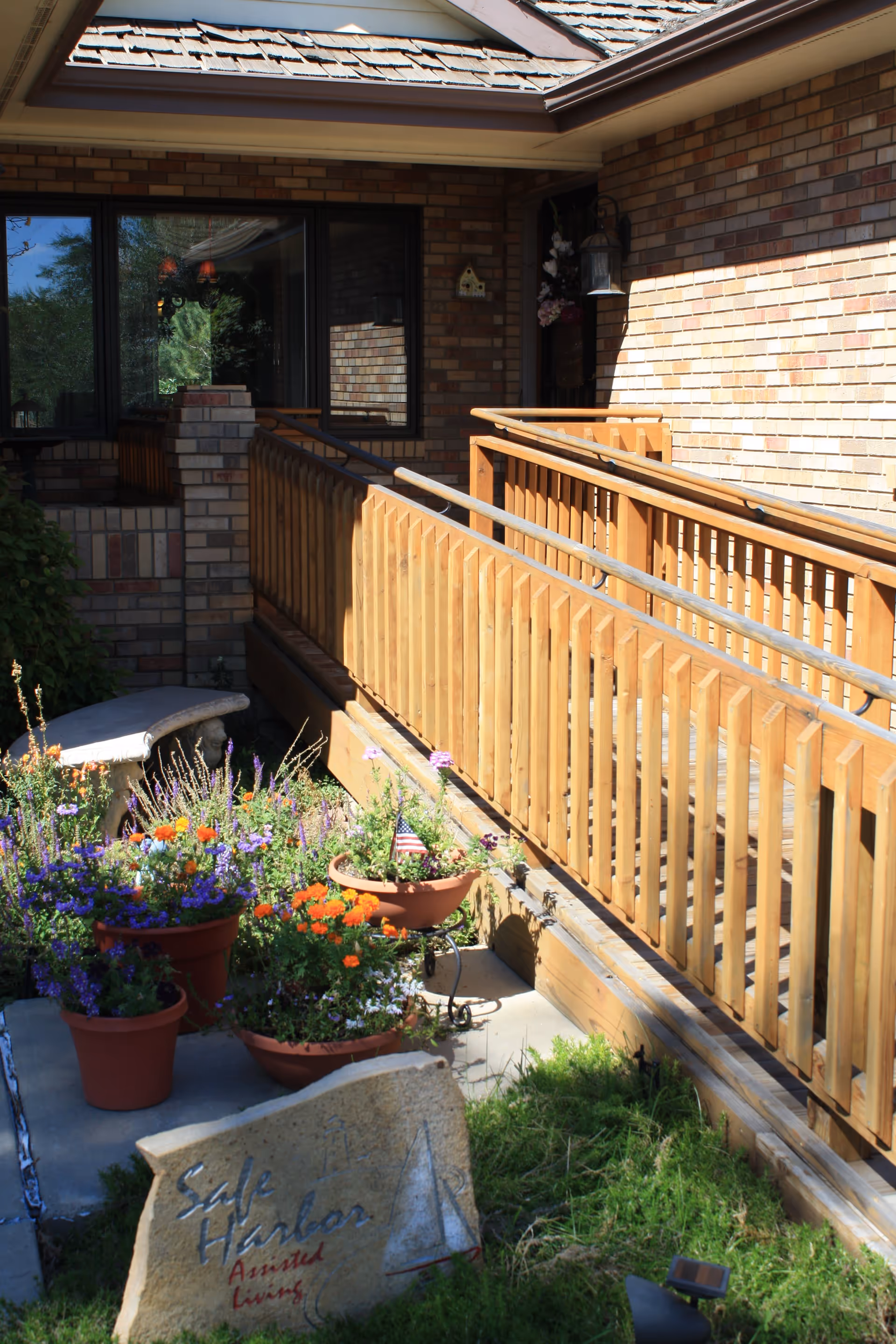 Outdoor view of a brick building entrance with a wooden ramp and handrails. There are several flower pots with colorful flowers and a stone sign that reads 'Safe Harbor Assisted Living'. A small American flag is placed in one of the flower pots.