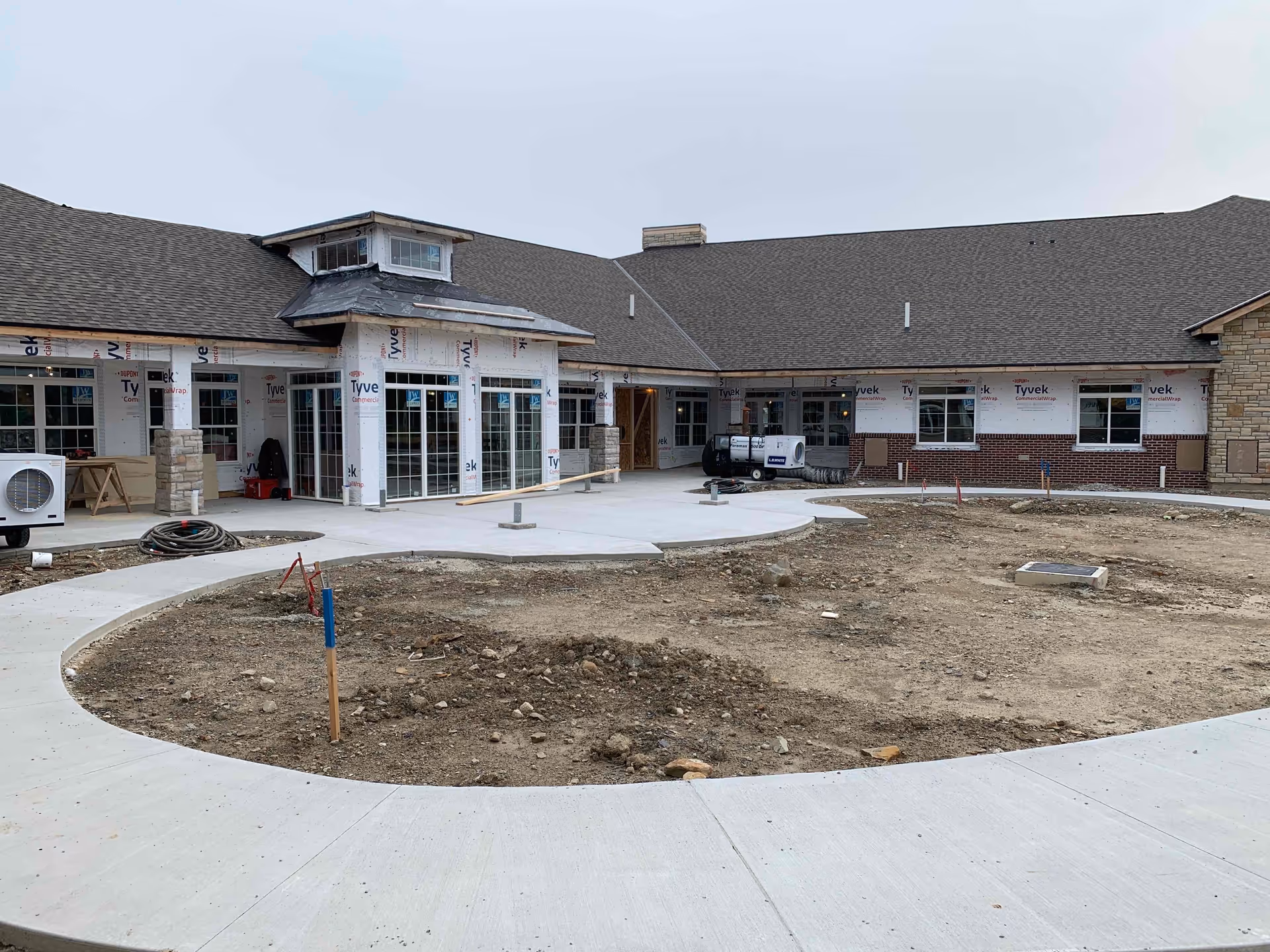 Exterior view of a building under construction with a large circular dirt area in the foreground surrounded by a concrete walkway. The building has a pitched roof and multiple windows, with some sections covered in Tyvek house wrap and others with brick and stone accents.