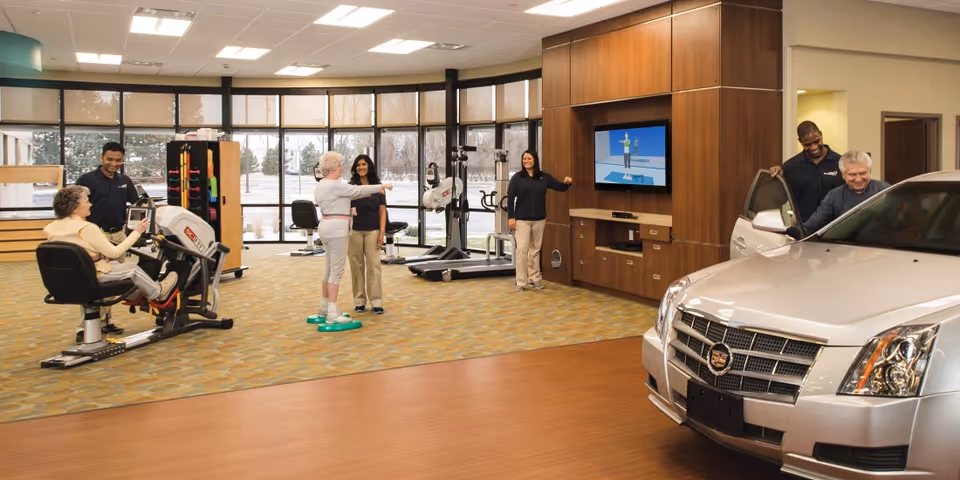 A rehabilitation center room with exercise equipment and a car inside. Two elderly individuals are engaged in physical therapy activities with the assistance of staff members. One person is using a seated exercise machine, another is balancing on a green platform while being guided, and a third person is getting into a silver Cadillac car with help from a staff member. The room has large windows and a TV screen displaying an exercise animation.