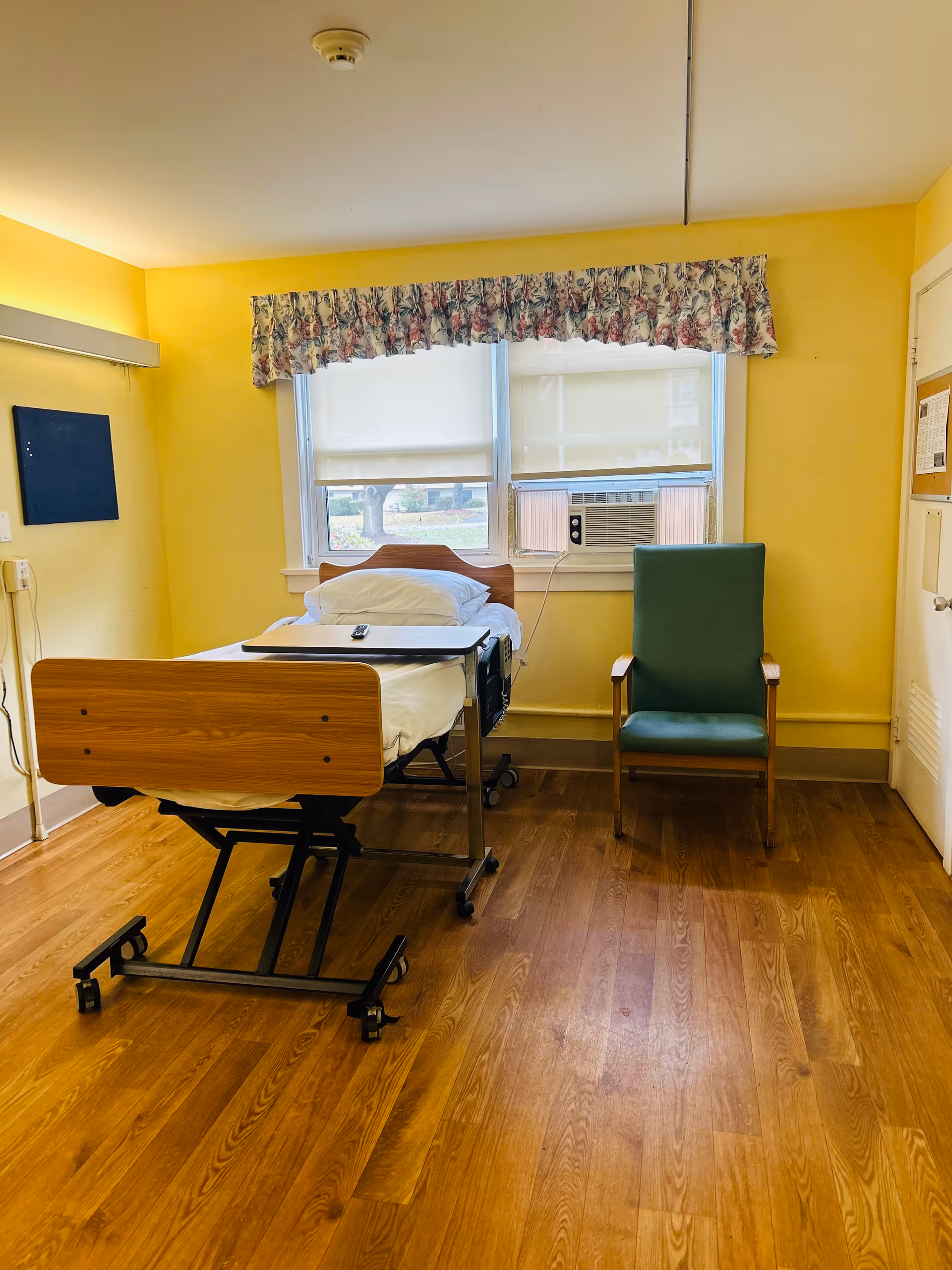 A small room with yellow walls and wooden flooring featuring a hospital-style bed with white bedding and a remote control on top. Next to the bed is a green cushioned chair with wooden armrests. The window behind the bed has floral curtains and an air conditioning unit installed.
