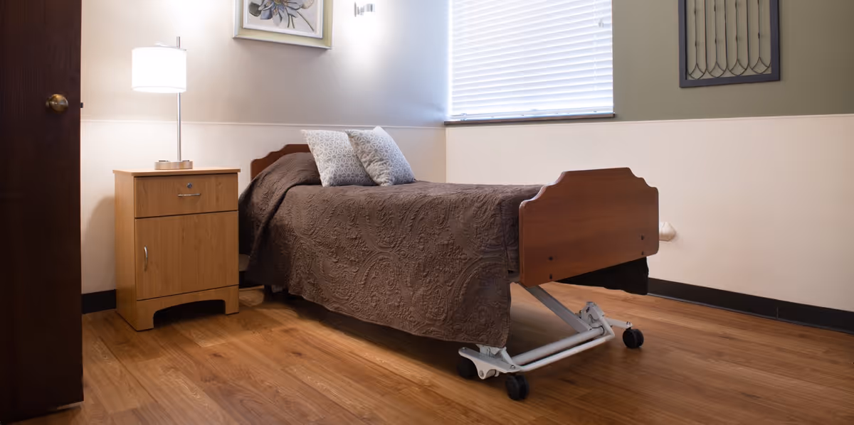 A simple senior bedroom with a single adjustable bed on casters, a wooden nightstand with a lamp, and a window with blinds.