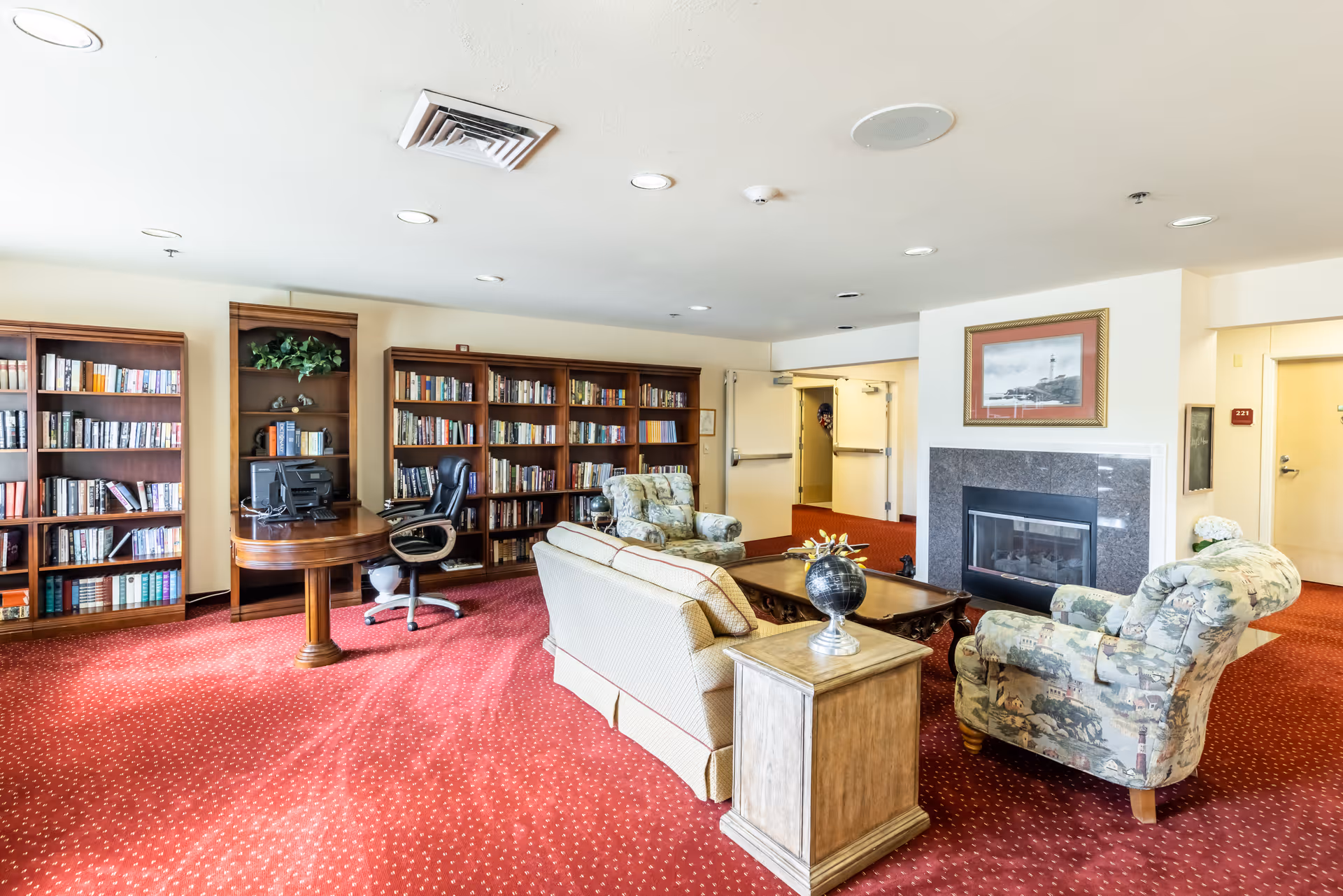 A cozy senior living common area with red carpet, a beige sofa, two patterned armchairs, a wooden coffee table, and a fireplace with a framed picture above it. There are multiple wooden bookshelves filled with books and a small desk with a computer and office chair. The room has white walls and ceiling with recessed lighting.