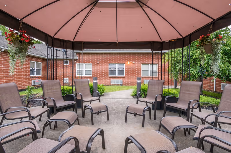 Covered outdoor courtyard with arranged lounge chairs and ottomans under a gazebo next to a brick building.