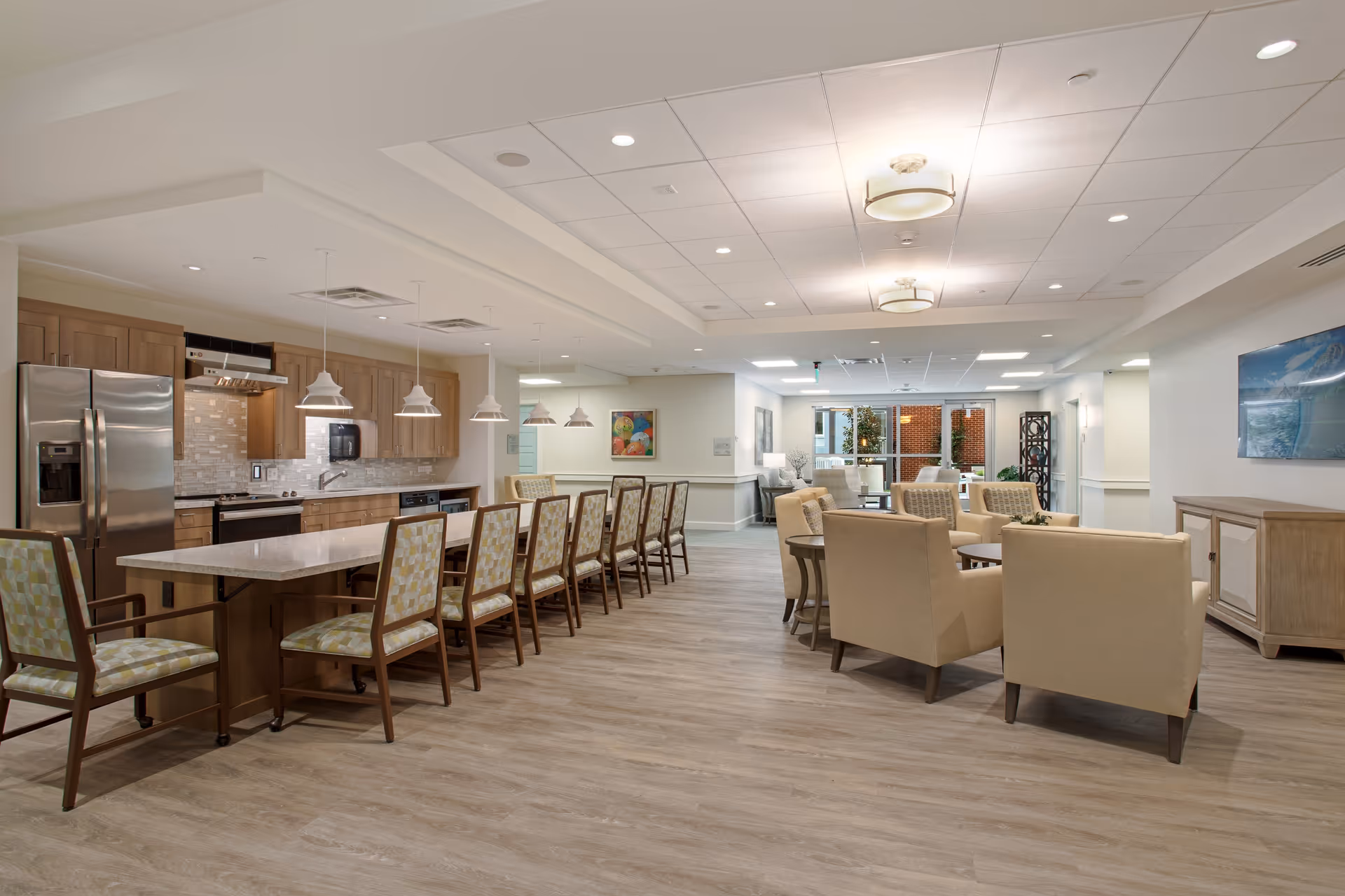 A spacious and modern common area in a senior living facility featuring a large kitchen island with multiple chairs, stainless steel appliances, pendant lighting, and a comfortable seating area with armchairs and tables. The room has wood flooring, recessed lighting, and large windows allowing natural light.